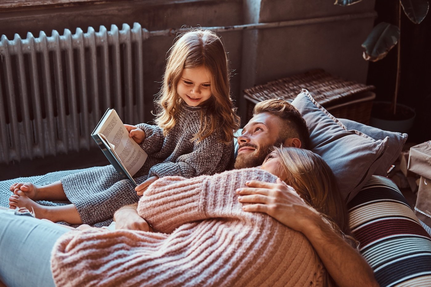 Family concept. Mom, dad and daughter reading storybook together while lying on bed.