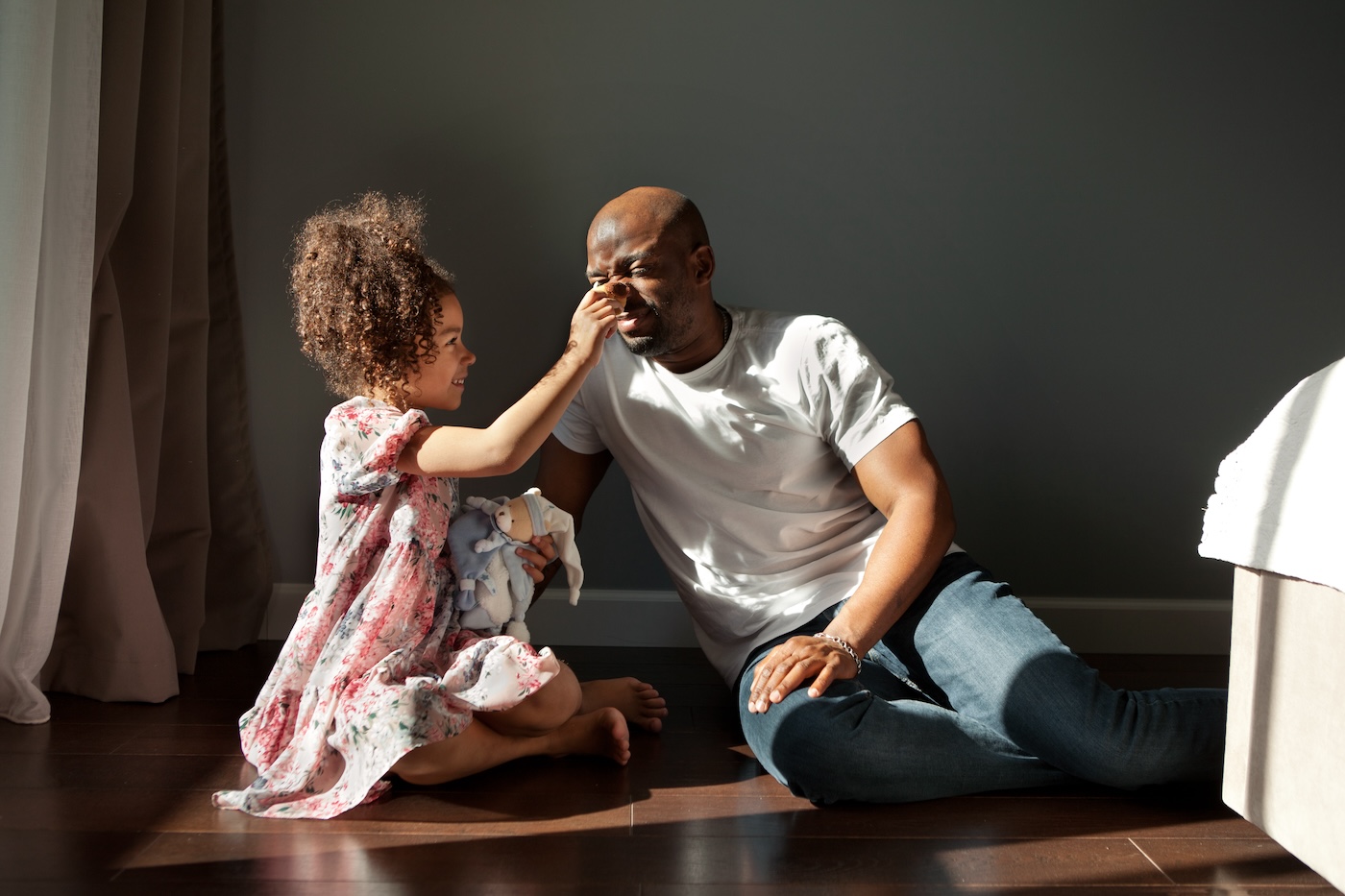 Happy father and his daughter playing with plush toy together. Child girl having fun with dad, sitting on floor at home, smiling. Authentic portrait on sunny day.