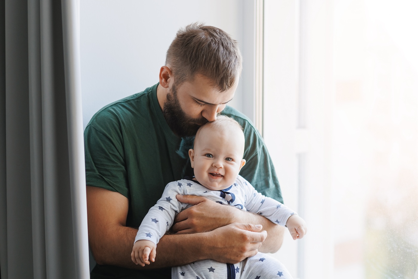 Father hugs baby boy, young kisses his son on head in living room. Concept lifestyle parenting fatherhood moment.