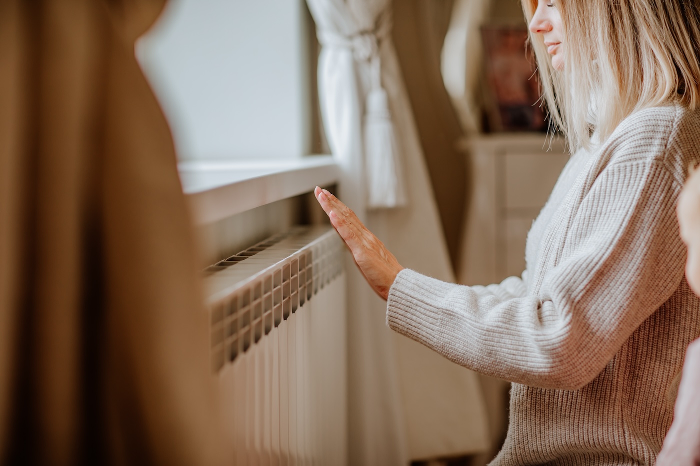 Young blond woman in long winter beige sweater is posing at home near the radiator. Winter season concept. Focus is at hands. Economy program